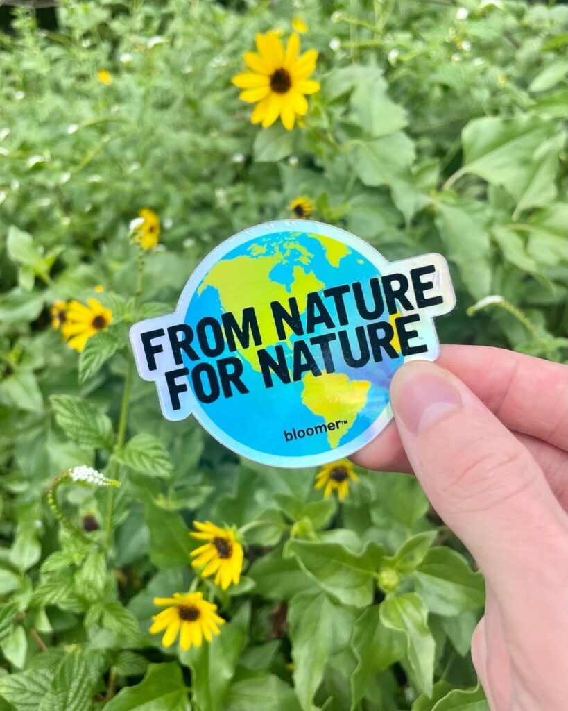 Hand holding a 'From Nature, For Nature' bloomer sticker with an Earth design, against a background of green plants and yellow flowers.