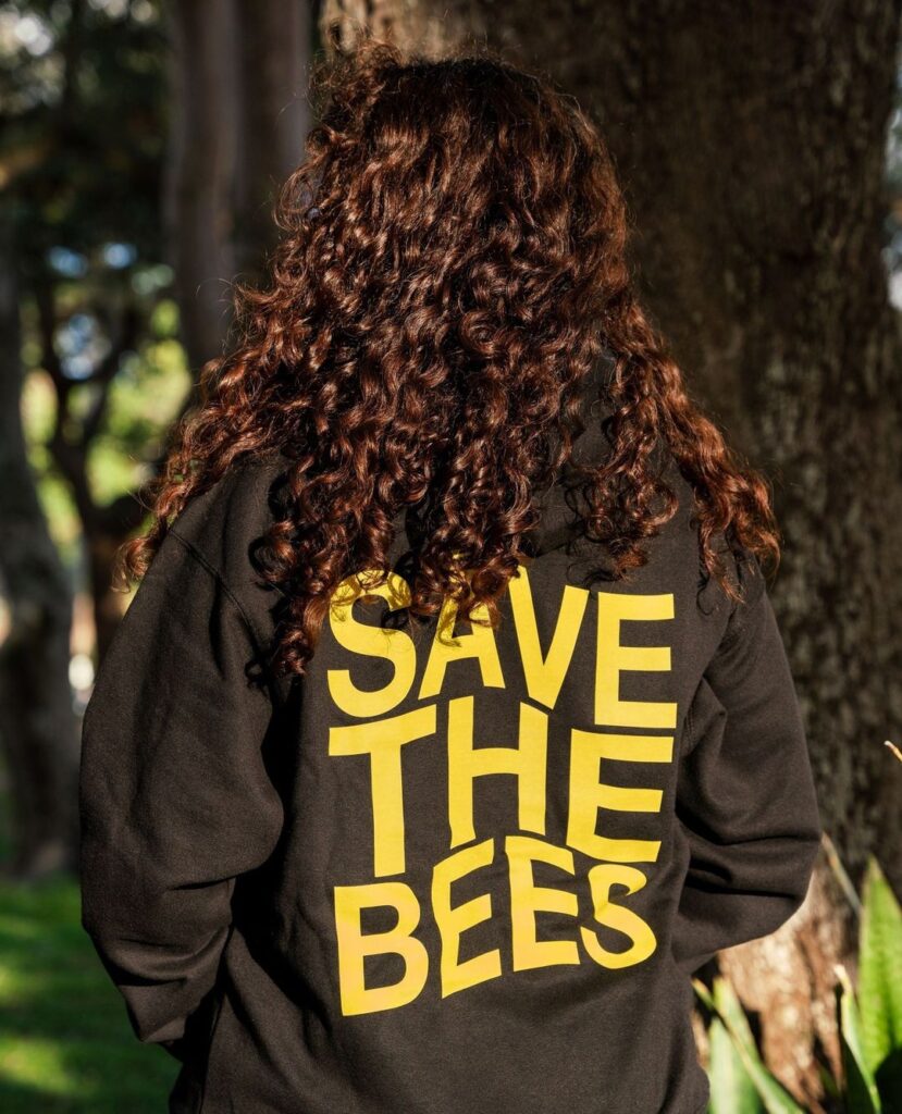 Person with long curly hair wearing a black sweatshirt featuring yellow text 'SAVE THE BEES,' promoting awareness for bee conservation.