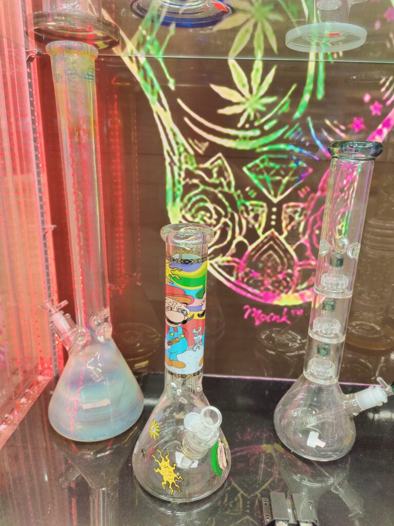 3 Bongs in a glass display, Featuring a Stoned Mario Bong, A glass bong with 4 percs and a bong with a tall bong with a blue hue bottom