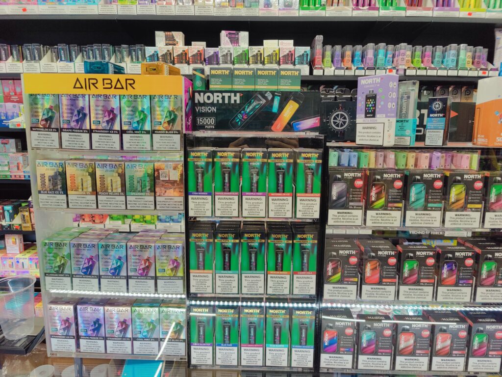 The smokeshop features displays of various vape products neatly arranged on shelves. Brands like Air Bar and North stand out with their colorful packaging, showcasing a wide array of flavors. The shelves are well-stocked, reflecting the shop's dedication to providing a diverse selection for vape enthusiasts. 