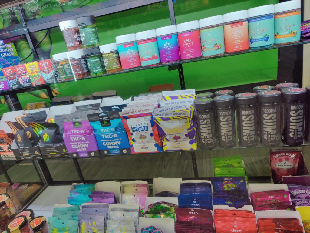 Shelves displaying assorted cannabis edibles and products in colorful packaging. SUMO Gummies, STIIIIZY, GUMMY CUBES, URB, DOZO, JUSTCBD, DIAMOND SHRUUMZ, CUREVANA, DOZO, SPACE DROPS, +MORE!