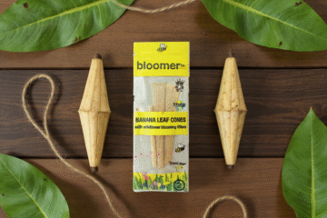 A box of Bloomer banana leaf cones with wildflower blooming filters is surrounded by green banana leaves, two wooden cones in shape of a banana, and twine on a nice wooden surface.
